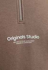 Ruskopparin vetonopean yliveto, joka on valmistettu sileästä kankaasta. Varustettu hopeanvärisellä vetoketjulla ja valkoisella tekstillä: "Originals Studio" pienemmän tekstin yläpuolella.