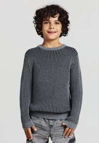 Maglione grigio a coste con maniche lunghe e scollatura rotonda, caratterizzato da strisce testurizzate, indossato sopra pantaloni di jeans grigi.
