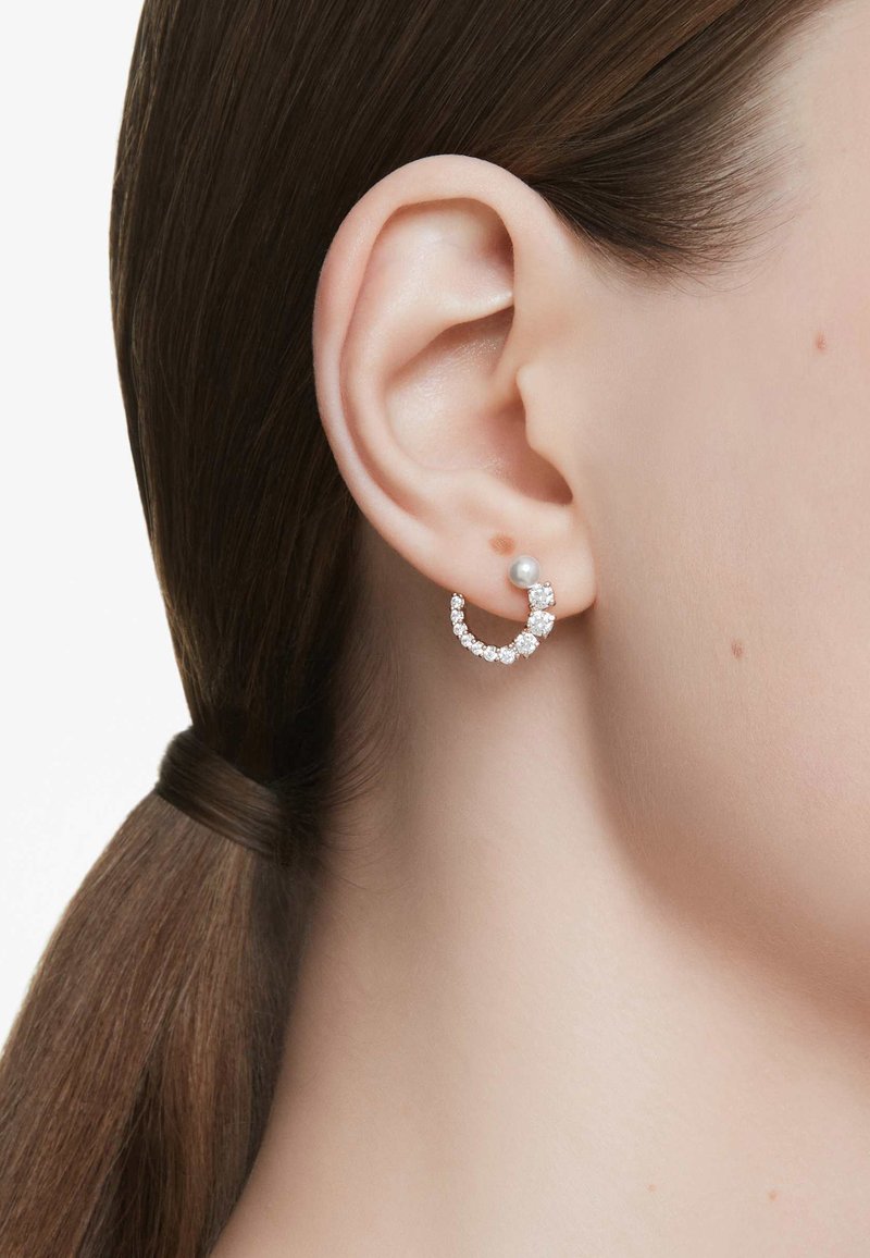 Strieborná kruhová náušnica s priehľadnými kryštálmi a perlovým akcentom, s zakriveným dizajnom, ktorý jemne obopína ušný lalôčik.