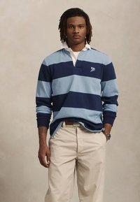 Jonge man met middelgrote dreadlocks, gekleed in een blauw gestreept rugbyshirt en beige broek, staand met één hand in zijn zak.