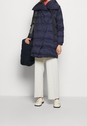 Manteau matelassé bleu marine avec un grand col, un design matelassé horizontal et des poignets élastiques. Associé à un pantalon large blanc et des mocassins crème.
