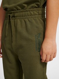 Close-up of a person wearing olive green sweatpants with elastic waistband, drawstring, and "hummel" logo on the upper thigh.