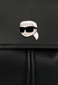 Musta nahkaisten käsilaukku, jossa on etuosassa sarjakuvamaistyylinen hopeapinssi, jossa on valkoiset hiukset ja mustat aurinkolasit päällään.