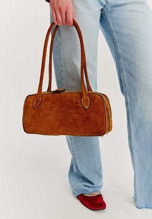 Osoba trzymająca prostokątną brązową zamszową torebkę, ubrana w jasnoniebieskie dżinsy i czerwone buty na ostro zakończonym nosku, stojąca na białej podłodze.