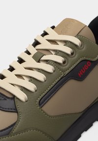 Κοντινό πλάνο ενός αθλητικού παπουτσιού σε μπεζ και ελιά χρώμα με κρεμ κορδόνια και κόκκινο λογότυπο "HUGO" στο πλάι.