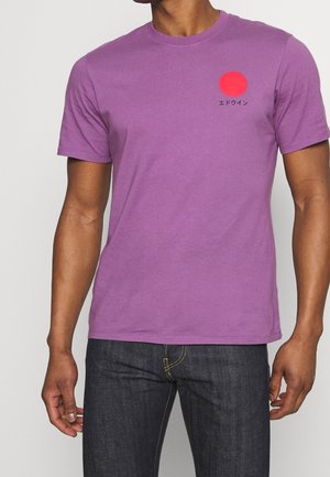 Mężczyzna ubrany w fioletowy t-shirt z krótkim rękawem z czerwonym kołem i japońskim tekstem na lewej piersi, w połączeniu z ciemnoniebieskimi dżinsami.