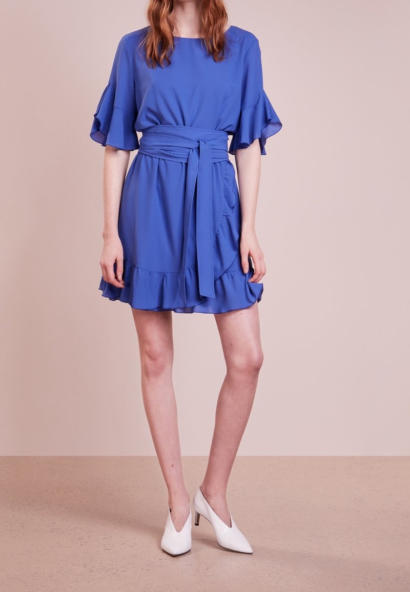 Femme portant une robe bleue à volants avec une large ceinture nouée à la taille et des chaussures blanches à bouts pointus et talons, debout sur un sol beige.