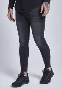 Osoba v černých skinny džínách a černých teniskách, s viditelnými tetováními na rukou a kotnících, stojící na světlém pozadí.