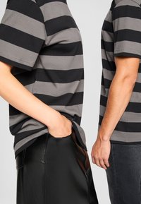 Črne in sive črtaste bombažne majice z gladko teksturo. Ena majica je vtaknjena v sijočo črno usnjeno krilo, medtem ko je druga v kombinaciji z denim hlačami.