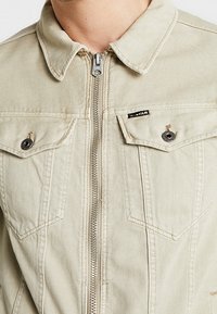 Beżowa kurtka jeansowa zapinana na zamek, z klapami na piersi, detalami na guziki oraz małą czarną metką z logo G-Star na lewym kieszeni.