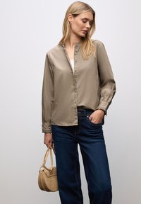 Blusa a maniche lunghe di colore taupe con chiusura a bottoni, vestibilità morbida e polsini arricciati, abbinata a jeans dritti blu scuro e una piccola borsa beige.
