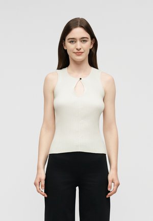 Fiatal nő egyenes barna hajjal, ujjatlan bordázott fehér felsőt visel kulcslyuk részlettel és fekete nadrágot, egyszínű háttér előtt.