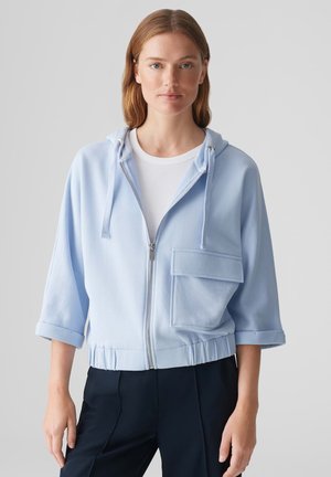 Lichtblauwe hoodie met rits, voorzien van een trekkoordkap, een voorzak en een elastische tailleband. Zachte stof met opgerolde mouwen en een ontspannen pasvorm.