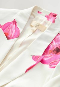 Leinenblazer mit großen rosa Blumenmotiven auf den Schultern und Taschen, in einer cremefarbenen Grundfarbe und mit glatter Textur.