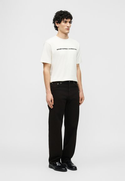 Άνδρας μοντέλο που φοράει λευκό t-shirt Emporio Armani, μαύρο παντελόνι και μαύρα παπούτσια, στέκεται μπροστά σε απλό ανοιχτό γκρι φόντο.