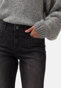 Svarta jeans med en åtsittande design, prydda med rhinestone-accenter på framsidan, i kombination med en ljusgrå stickad tröja.