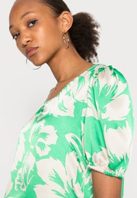 Blusa floral verde com mangas bufantes, apresentando padrões florais brancos. Feita de um tecido suave e texturizado, com um decote amplo e acabamentos elásticos.
