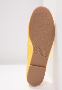 Semelle d'une chaussure jaune indiquant le nom de la marque Tamaris et la taille 37, avec une semelle en caoutchouc marron texturée présentant des motifs en forme de W.