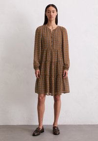 Braun gemustertes Kleid mit langen Ärmeln, das ein gestuftes Design und einen Knopfverschluss aufweist. Akzente setzen weiße geometrische Formen im gesamten Kleid.