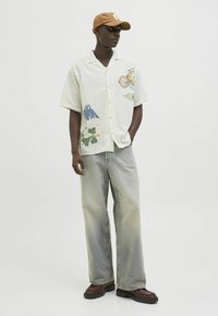 Short-sleeve light green shirt with floral print, paired with light-wash wide-leg jeans and brown chunky shoes. The model wears a brown cap.