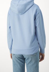 Světle modrá mikina s kapucí na šňůrku, jemná textura, žebrované manžety a volný střih, v kombinaci se světle modrými džíny.