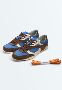 Baskets en daim bleu et marron avec des lacets marron foncé, accompagnées d'un lot de lacets orange étiquetés Oysho à côté.
