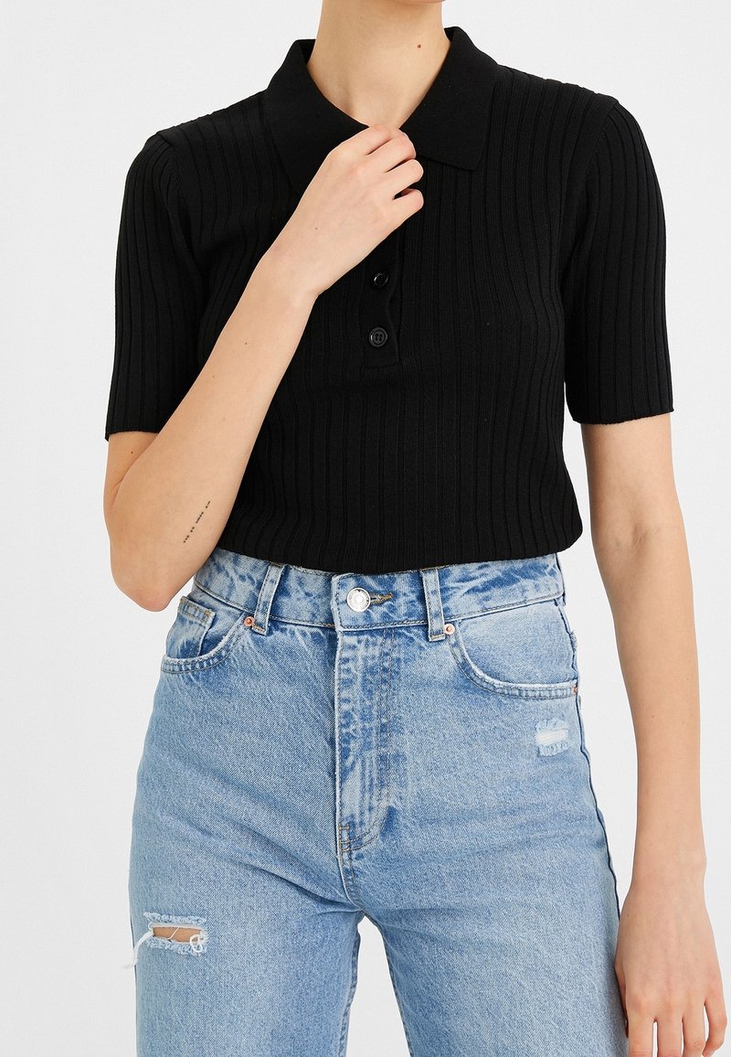 Personne portant un polo noir côtelé à manches courtes et un jean bleu clair déchiré taille haute, main touchant le col.