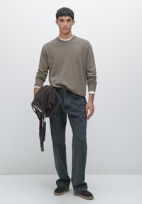 Bruine trui met geribbelde manchetten, donkergrijze rechte spijkerbroek en brune schoenen. Model houdt een bruine tas met een schouderband vast.