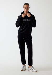 Vrouw in een zwarte velours hoodie en broek met witte sneakers, staande en haar hoodie-kraag aanpassend tegen een effen achtergrond.