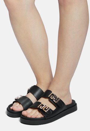Black slide sandals with two wide straps, featuring metallic gold buckles. Thick sole with textured tread for grip. Smooth leather material.