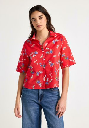 Ung kvinde iført rød kortærmet skjorte med små blå og hvide blomsterprint og blå jeans, stående mod en ensfarvet baggrund.