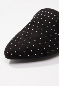 Svart mockaskor med spetsig tå, prydda med silverfärgade nitar som är ordnade i ett prickigt mönster över den övre ytan.