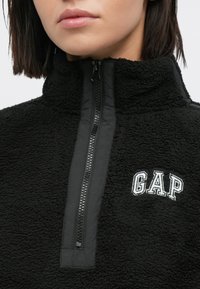 Pullover in pile nero con chiusura a zip, collo alto e logo "GAP" ricamato. Tessuto testurizzato con accenti lisci lungo la zip.