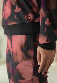 Close-up of a hand resting beside floral-patterned textured pants and matching top with black cuffs and waistband, featuring a small gold charm.