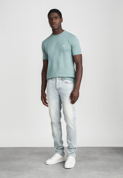 Svijetlozelena majica s kratkim rukavima i okruglim izrezom, u kombinaciji s svijetloplavim izblijedjelim trapericama i bijelim tenisicama. Minimalno brendiranje na majici.