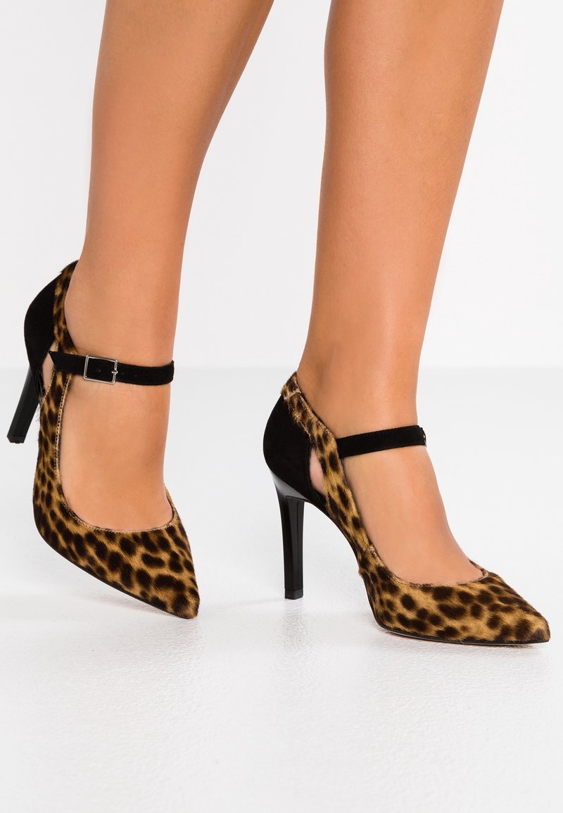 Jambes portant des talons hauts imprimés léopard avec des pointes pointues et des brides noires à la cheville sur un fond blanc.