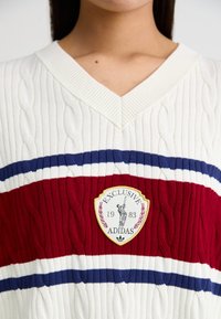 Pulover s kabelskim pletenjem v kremni barvi, ki ima rdeče in modre črte. Oblika v vratu v obliki črke V z logotipom, ki prikazuje "EXCLUSIVE 1983 ADIDAS."