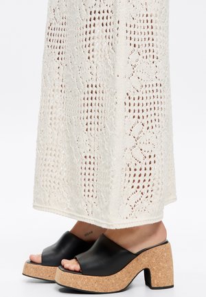 Sandali wedge neri in pelle con piattaforma in sughero, dotati di punta aperta e superficie liscia. Visti con un indumento bianco lavorato a maglia sullo sfondo.