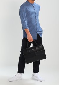 Valigetta nera in pelle con due tasche frontali e manici doppi, abbinata a una camicia blu a bottoni e pantaloni neri. Sneakers bianche indossate.