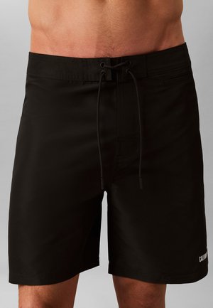 Czarne kąpielówki męskie z talią na sznurek i małym logo na lewej nogawce, noszone przez osobę bez koszulki od talii do połowy uda.