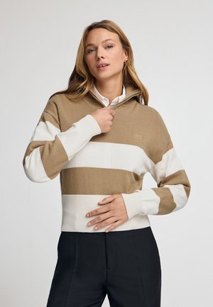 Beige-weißer gestreifter Pullover mit Reißverschlusskragen und gerippten Bündchen, kombiniert mit dunklen Hosen. Stylische Stricktextur.