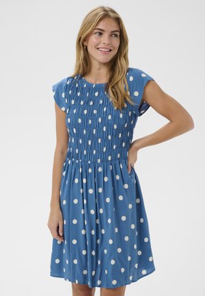 Mujer sonriente con cabello rubio, vestida con un vestido azul de manga corta con lunares blancos, de pie con una mano en la cadera.