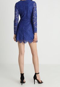 Robe en dentelle bleue à manches longues, ourlet festonné et silhouette ajustée. Portée avec des talons à brides cheville noirs.