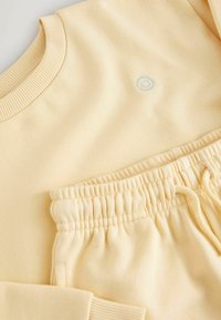Sudadera amarilla con cuello acanalado y pequeño logo de sonrisa azul; pantalones joggers a juego con cintura elástica y cordón. Textura suave de algodón.