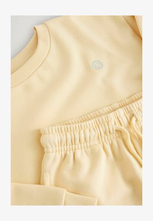 Gul sweatshirt med ribbet krave og lille blå smiley-logo; matchende joggers med elastisk taljebånd og snøre. Blød bomuldstekstur.