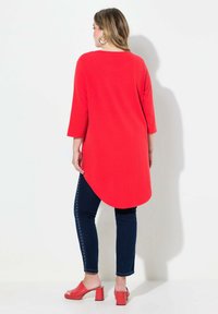 Tunica lunga rossa con orlo curvo, abbinata a jeans blu scuro con borchie laterali e mule con tacco blocco rosso. Elementi di design semplici.