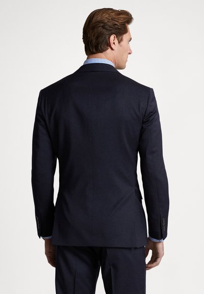 Tamsiai mėlynas kostiumo švarkas su struktūruotais pečiais, vienu šonu ir užsegamais rankogaliais. Po juo matyti šviesiai mėlynas languotas marškinis.