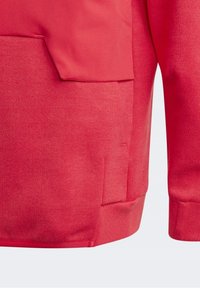 Felpa rossa con tessuto testurizzato, tasca a marsupio e polsini a costine. Il design include un dettaglio di tasca laterale.