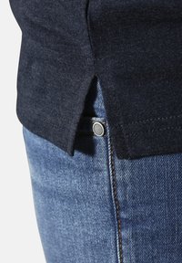 Dunkelblaues Baumwollhemd mit Seitenschlitz, kombiniert mit hellblauen Jeans aus Denim, die einen silbernen Knopf und orangefarbenen Nähten haben.