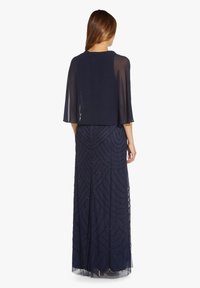 Žena oblečená v tmavě modré, dlouhé večerní šaty se zdobenými geometrickými vzory a průsvitným pláštěm přehozeným přes ramena, stojící s obratem k nám.